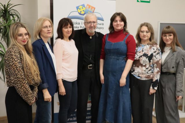 Na zdjęciu znajduje się sześć kobiet, a między nimi po środku stoi Krzysztof Daukszewicz, za nimi stoi baner z logiem biblioteki.
