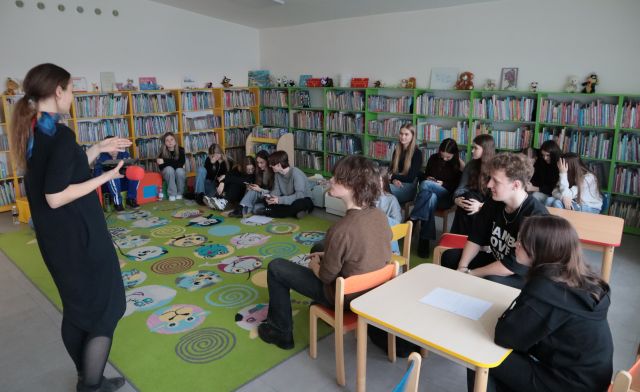 młodzież siedzi na dywanie, pufach i krzesełkach, przed nimi kobieta, w tle regały z książkami