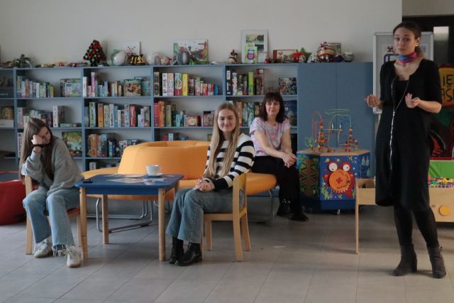 przy stoliku siedzą dwie dziewczyny, na pufie siedzi dyrektorka biblioteki, stoi kobieta