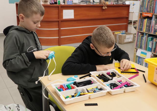 Na zdjęciu znajduję się dwójka chłopców, jeden siedzi przy stoliku, na którym leżą części lego i coś składa, a drugi stoi obok niego trzymając jeden element w ręce. Z tyłu znajduję się biurko.