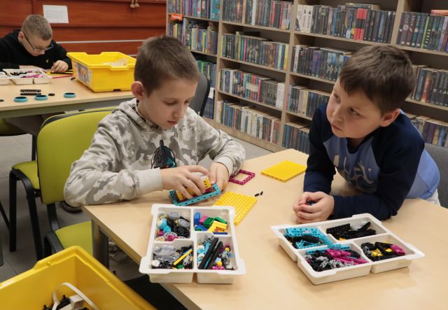 Na zdjęciu znajduję się dwójka chłopców przy stoliku i składają klocki Lego. W tle widać stolik przy którym ktoś siedzi oraz półki z książkami.