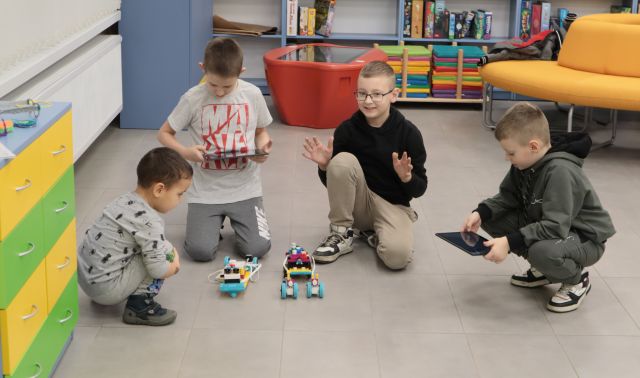 Na zdjęciu znajduje się czwórka chłopców dwoje na kolanach i dwójka kucająca. Dwóch z nich trzyma tablet, a przed nimi stoją wykonane przez nich robo-sanie z Lego. w tle widać półki szafę stolik interaktywny oraz kanapę.