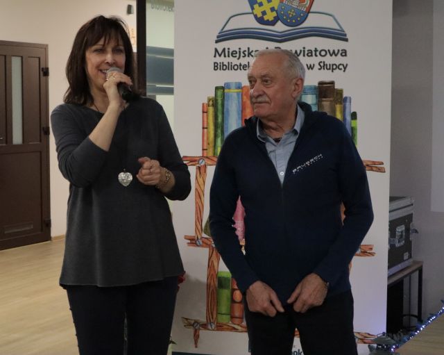 Na zdjęciu znajduję się kobieta z mikrofonem w ręku obok niej stoi mężczyzna. W tle znajduję się baner z książkami i logiem biblioteki.