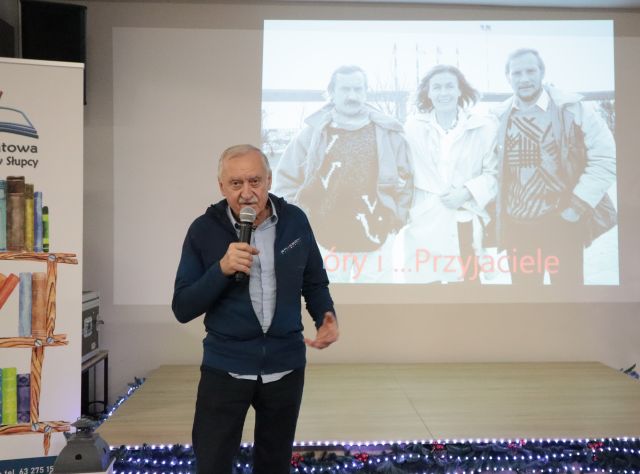 Na zdjęciu znajduje się Krzysztof Wielicki z mikrofonem w ręku. Z mężczyzną znajduje się prezentacja przedstawiająca trójkę ludzi, dwóch mężczyzn i kobietę w środku. Po lewej stronie znajduję się baner z książkami i logiem biblioteki.