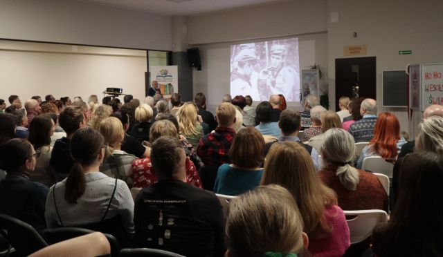 Na zdjęciu znajdują się ludzie skierowani w stronę sceny i wyświetlanej prezentacji przedstawiającej dwójkę ludzi.