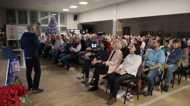 Na zdjęciu znajdują się siedzący ludzie skierowani w stronę mężczyzny z mikrofonem. W tle znajdują się okna oraz choinka.