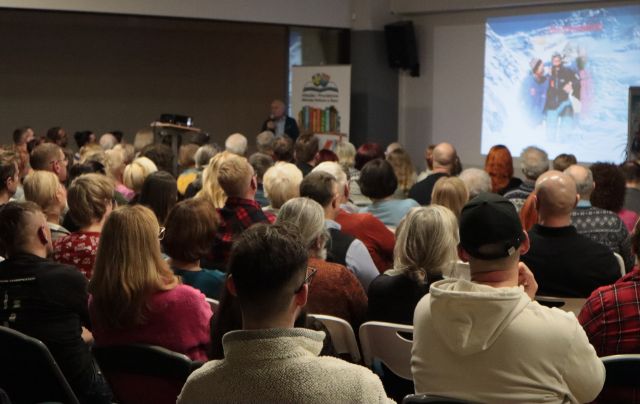 Na zdjęciu znajduję się widownia skierowana w stronę sceny przed którą stoi mężczyzna z mikrofonem. W tle znajduję się prezentacja przedstawiająca ludzi w górach oraz baner z książkami i logiem biblioteki.