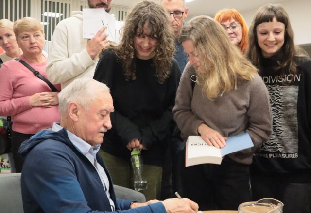 Na zdjęciu znajduję się siedzący bokiem przy stoliku mężczyzna a przy nim znajdują się ludzie w kolejce na zdjęcie i podpis książki.