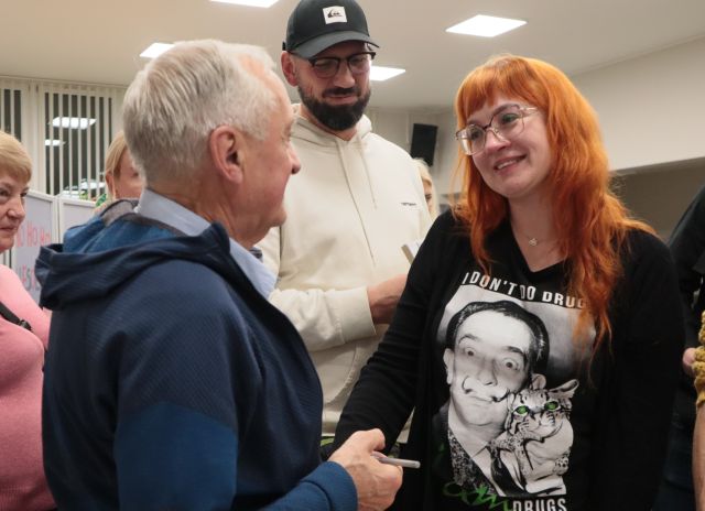 Na zdjęciu znajduję się mężczyzna oraz kobieta, którzy podają sobie rękę. Za nimi widoczny jest mężczyzna. Zdjęcie zostało zrobione od boku.