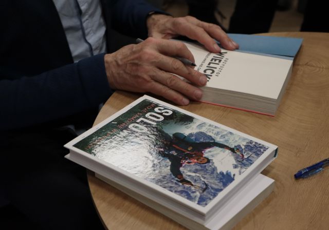 Na zdjęciu znajdują się dłonie mężczyzny trzymające długopis oraz książkę. Na stoliku znajduję się również książka okładką skierowaną ku górze.