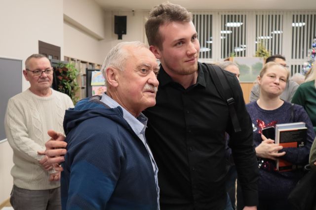 Na zdjęciu znajduję się dwójka mężczyzn pozujących do zdjęcia, w tle znajdują się ludzie czekający na podpis książki.