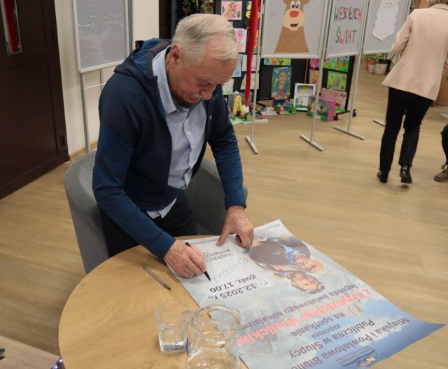 Na zdjęciu znajduję się Krzysztof Wielicki podpisujący plakat promujący spotkanie z nim. W tle znajduję się kobieta tyłem oraz wystawka prac plastycznych.