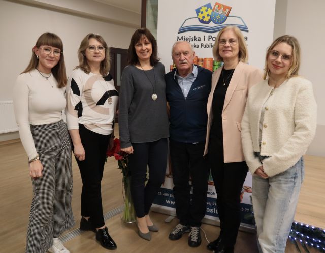 Na zdjęciu znajduję się Krzysztof Wielicki oraz pięć bibliotekarek, trzy stoją po lewej stronie, a dwie po prawej. Za nim znajduję się baner z logiem biblioteki.
