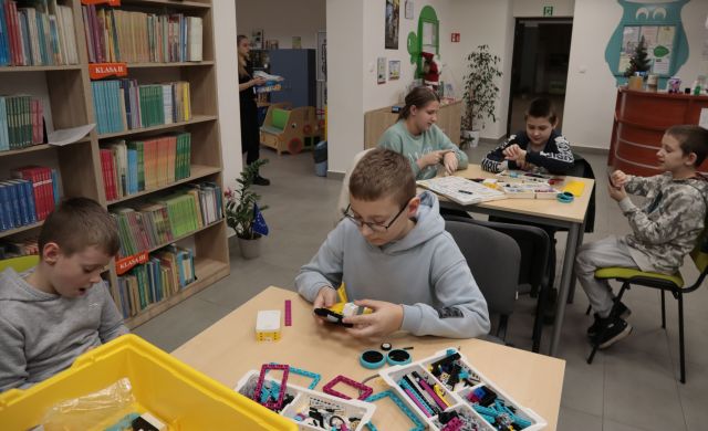 Na zdjęciu znajduję się dwójka chłopców przy stoliku składających LEGO, za nimi przy stoliku siedzi dwójka chłopców i jedna dziewczyna, którzy również układają LEGO. Po lewej stronie znajdują się półki z książkami.