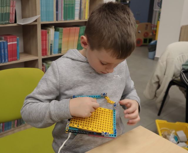 Na zdjęciu znajduję się chłopiec trzymający w rękach Lego, ma głowę skierowaną w dół. W tle znajdują się półki z książkami.