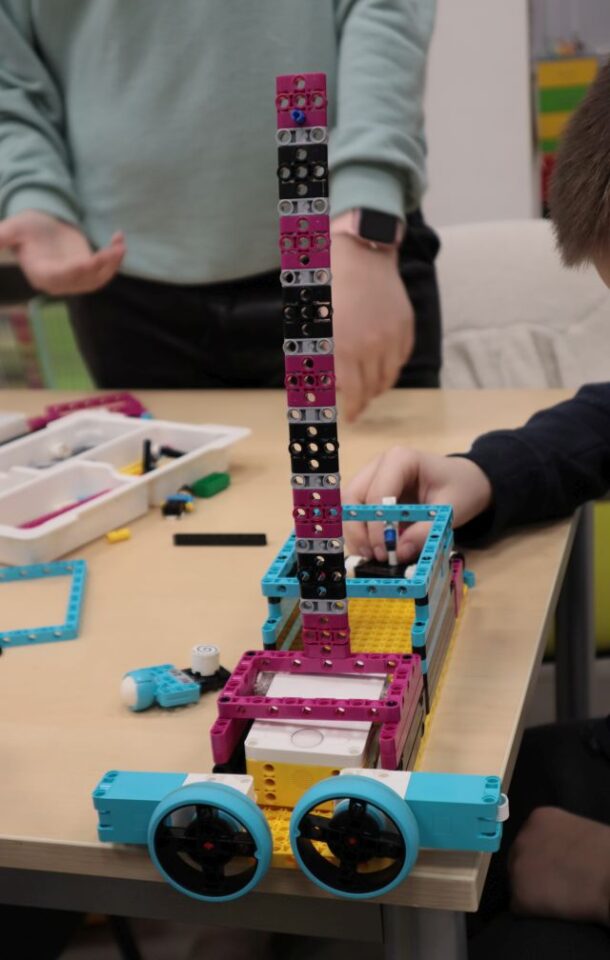 Na zdjęciu znajduję się zbudowany z klocków LEGO samochód z wysoką anteną również z klocków. Na stole znajdują się również klocki, a w tle widoczne są ręce dzieci.