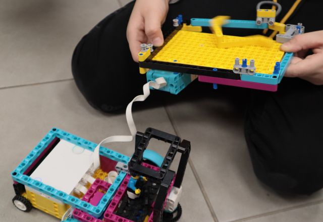 Na zdjęciu znajduję się chłopiec trzymający jedną część samochodu zbudowanego z LEGO w rękach, druga podłączona do niego część leży na ziemii.
