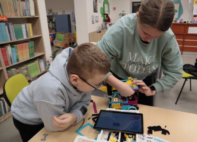 Na zdjęciu znajduję się chłopiec oraz stojąca koło niego dziewczyna. Chłopiec patrzy na tablet, a dziewczynka składa LEGO. W tle znajdują się półki z książkami.