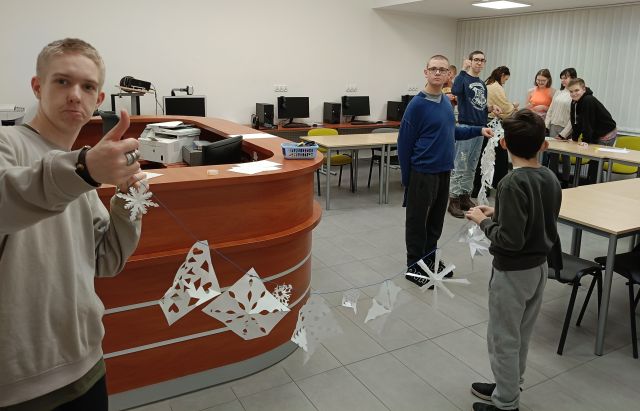 Na zdjęciu znajduje się młodzież, jeden chłopiec stoi po lewej stronie i trzyma za sznurek, reszta stoi po stronie prawej również trzymając za sznurek, na którym powieszone są śnieżynki z papieru. Młodzież znajduje się w sali komputerowej.