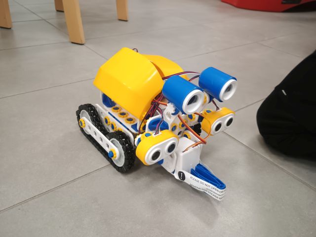 Na zdjęciu znajduje się robot Skribot, jeżdżący po podłodze, obok niego znajduje się noga chłopca.