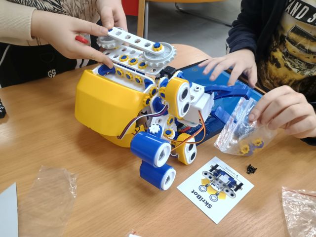Na zdjęciu znajduje się robot na stoliku oraz dwóch chłopców, którzy go budują.