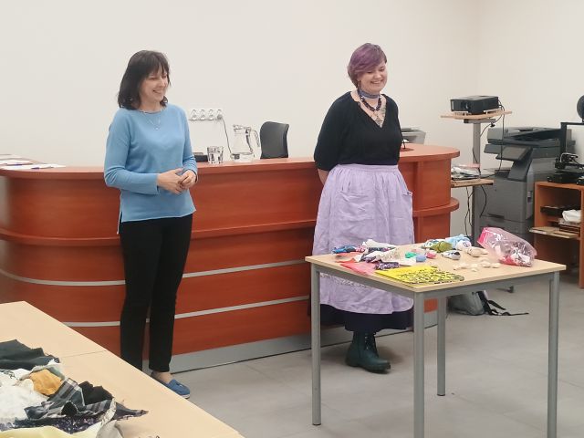 Na zdjęciu znajdują ię dwie kobiety, jedna stoi przed stolikiem, na którym leżą materiały. Za kobietami znajduje się biurko biurowe. 