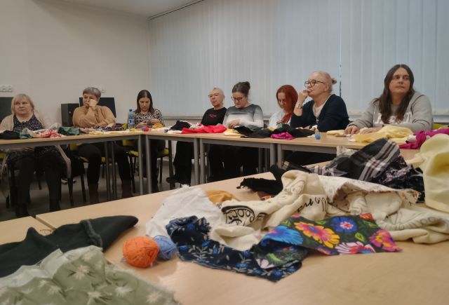 Na zdjęciu znajdują się stoły ułożone w literę C, na których leżą kolorowe materiały, przy stolach siedzą kobiety i wykonują lalki. W tle znajdują się okna.