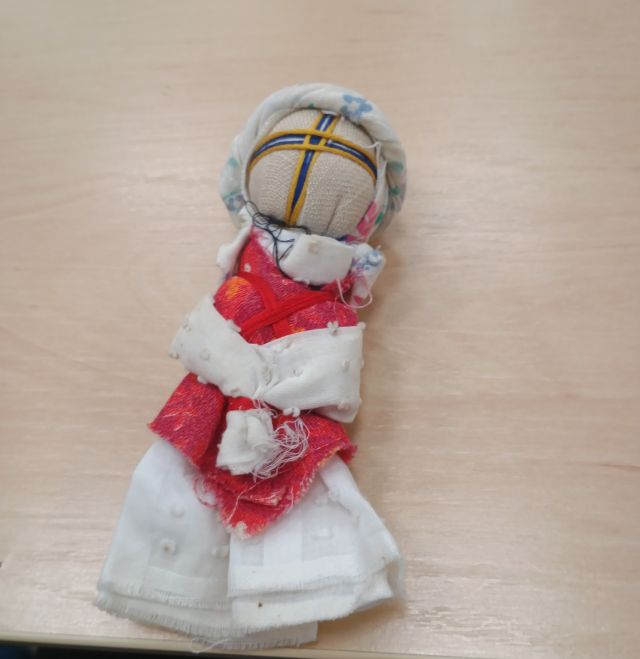 Na zdjęciu znajduje się motana lalka, w białej i czerwonej chuście i krzyżem na twarzy.