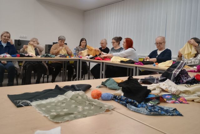 Na zdjęciu w centrum znajdują się połączone stoły, na których znajdują się kolorowe materiały, w tle znajdują się kobiety przy stole wykonujące lalki motane. Zajęcia odbywają się w sali komputerowej.