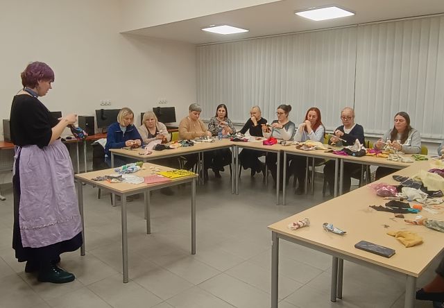 Na zdjęciu znajdują się kobiety przy stołach złączonych w literę C robiące ze znajdujących się na stole materiałów motane lalki. Przed nimi stoi kobieta przed stolikiem i pokazuje jak je wykonać. Warsztaty odbywają się w sali komputerowej i w tle znajdują się okna.