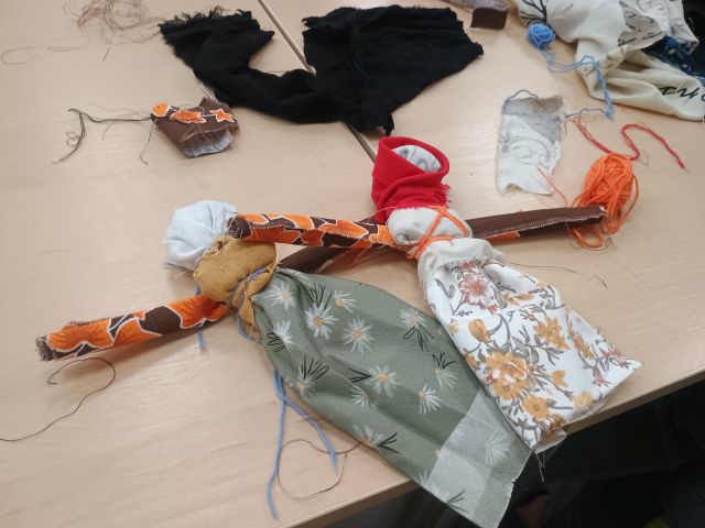 Na zdjęciu znajdują się dwie motane lalki położone na stole koło kolorowych materiałów.