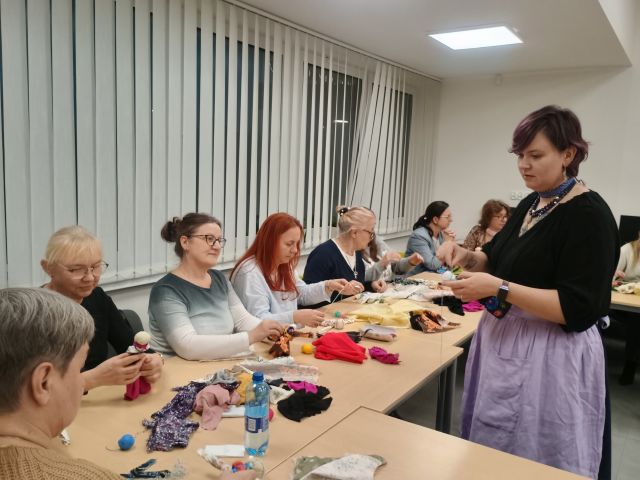 Na zdjęciu znajdują się kobiety przy złączonym stole i patrzą na kobietę po drugiej stronie stołu, która im pokazuje jak wykonać motane lalki z materiałów, która leżą na stole. W tle znajdują się okna.