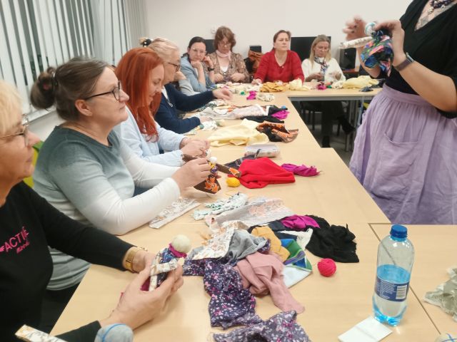 Na zdjęciu znajdują się kobiety przy złączonych stołach kończące wykonane przez siebie motane lalki z materiałów dostępnych na stole, po drugiej stronie stołu stoi kobieta i pokazuje kobietą jak je wykonać. Zajęcia odbywają się w sali komputerowej.