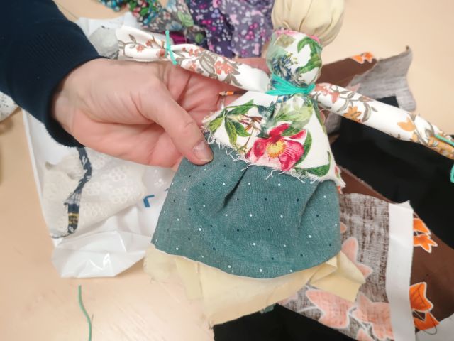 Na zdjęciu znajduje się kobieca ręka trzymająca wykonaną przez siebie motaną lalkę, która znajduje się na tle kolorowych materiałów.