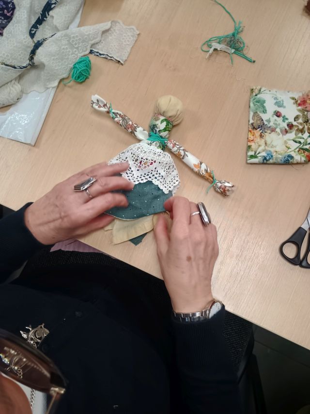 Na zdjęciu znajdują się kobiece ręce poprawiające spódnice wykonanej przez siebie motanej lalce. Dookoła znajdują się resztki materiałów.