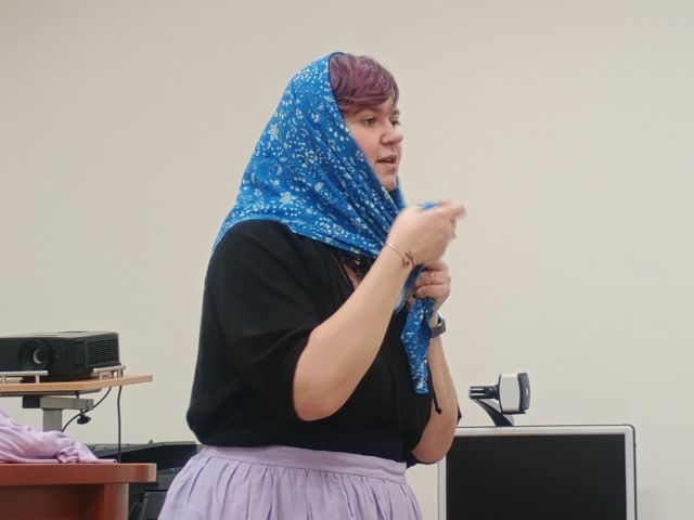 Na zdjęciu znajduje się kobieta stojąca bokiem pokazująca jak zawiązać chustę na głowie. Kobieta znajduje się w sali komputerowej.