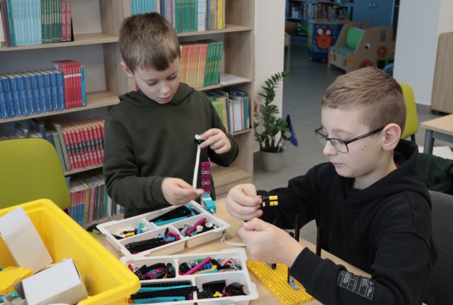 Na zdjęciu znajduję się dwójka chłopców siedzących przy stoliku i budująca z klocków LEGO. W tle znajdują się półki z książkami.