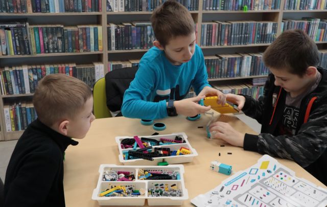 Na zdjęciu znajduję się trzech chłopców przy stoliku, którzy budują z LEGO, na stoliku leży reszta klocków. W tle znajdują się półki z książkami.