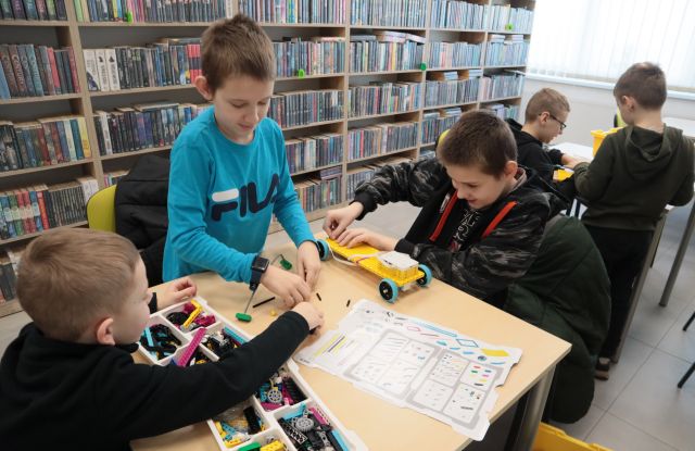 Na zdjęciu znajduję się trzech chłopców przy stoliku składających samochód z LEGO education, za nimi przy stole siedzi dwóch chłopców, którzy również budują z LEGO. W tle znajdują się półki z książkami.