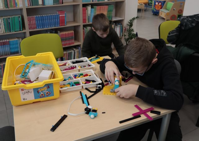 Na zdjęciu znajduję się dwójka chłopców przy stole, na którym znajdują się elementy z LEGO Education. Jeden chłopiec składa, a drugi znajduję się obok niego. W tle znajdują się półki z książkami.