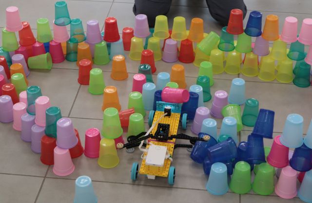 Na zdjęciu znajduję się konstrukcja z LEGO przypominająca długi samochód, wokół niej znajdują się kolorowe kubki, które pcha do przodu.