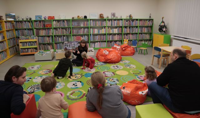 Na zdjęciu znajduję się kobieta, która siedzi na dywanie i pokazuje książkę dzieciom i rodzicom siedzącym na przeciwko niej. W tle znajdują się półki z książkami oraz dwie czerwone pufy do siedzenia.