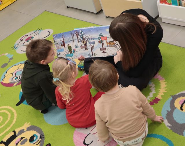 Na zdjęciu znajduję się kobieta, która trzyma otwartą książkę przedstawiającą zimowy krajobraz, obok siedzi trójka dzieci i przygląda się ilustracji. Zdjęcie zostało wykonane od góry.