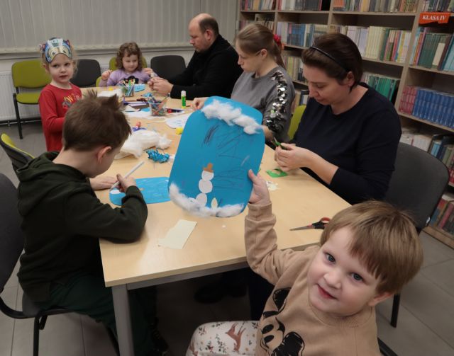 Na zdjęciu znajduję się dziewczynka przy stole, która trzyma w ręce wykonany przez siebie zimowy krajobraz. W tle przy stoliku znajdują się rodzice wraz z dziećmi, którzy wykonują prace plastyczne z kolorowych kartek i kredek, które znajdują się na stole.