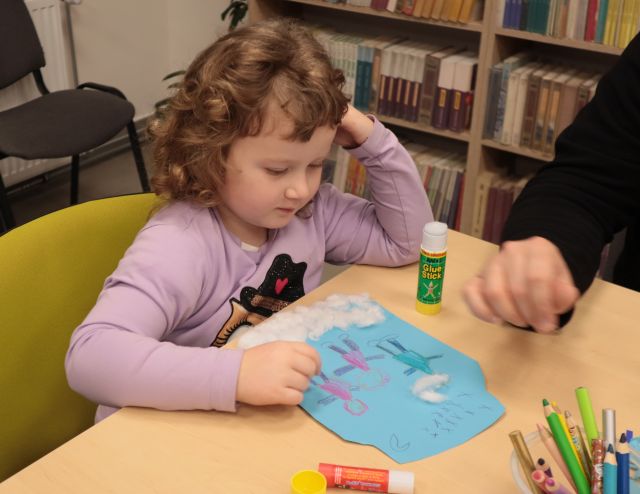 Na zdjęciu znajduję się dziewczynka rysująca na niebieskiej kartce, koło niej na stoliku znajduję się męska ręka. W tle znajdują się półki z książkami.