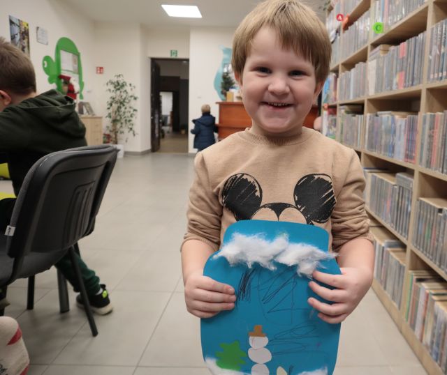 Na zdjęciu znajduję się dziewczynka trzymająca przed sobą wycięty papierowy słoik z niebieskiej kartki, na którym narysowany jest zimowy krajobraz. Po prawej stronie znajdują się półki z książkami, a po lewej stronie krzesełka.