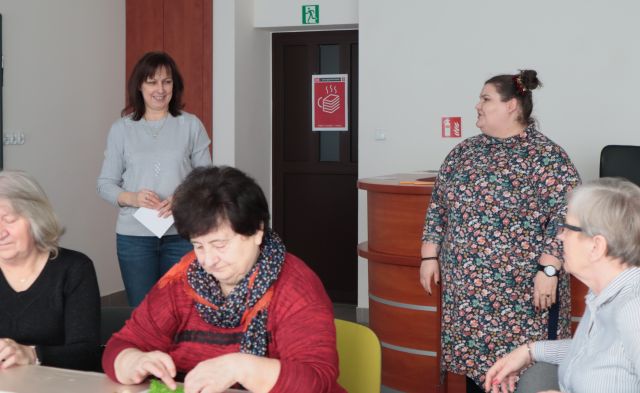 Na zdjęciu znajduję się pięć kobiet, trzy siedzą przy stole, a dwie stoją za nimi.