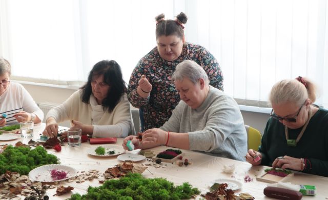 Na zdjęciu znajduje się pięć kobiet, cztery siedzą przy stole i robią obrazy z mchu, jedna stoi za nimi i im coś tłumaczy. W tle znajdują się okna.
