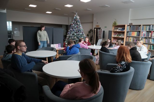 Na zdjęciu znajduję się trzynaście osób, które siedzą przy stolikach w czytelni i patrzą w kierunku stojącej kobiety. W tle znajdują się półki z książkami oraz choinka.