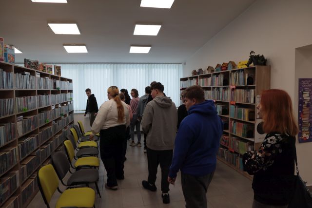 Na zdjęciu znajduję się młodzież, która idzie między półkami z książkami, przy regałach stoją krzesła. W tle znajdują się okna.